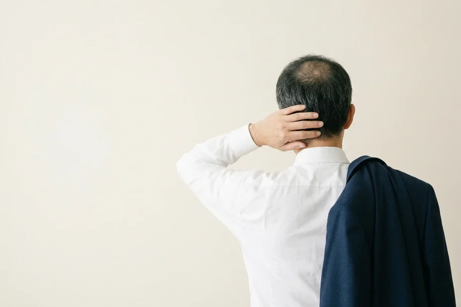 朝、上着を肩にかけ、後頭部に手をやる白いワイシャツの男性の後ろ姿。はじめの一歩は、医師に相談すること。